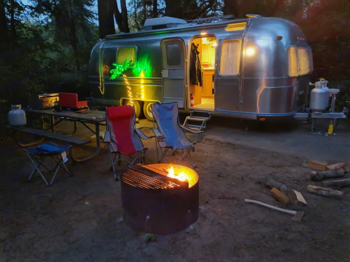 Cozy rv rental camping setup with Airstream, glowing campfire, and chairs at dusk. Perfect outdoor adventure.. Airstream Tradewind 1979