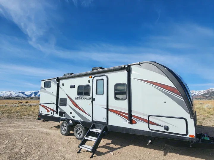 A white RV rental, a travel trailer with steps down, is set in a vast landscape with distant snow-capped mountains.. Heartland Wilderness 2019