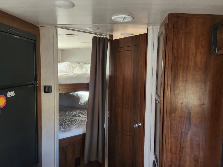 Inside an RV rental: a compact living space featuring a Dometic fridge, bunk beds with white linens, and a private wood door.. Heartland Wilderness 2019