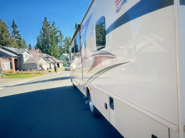 An RV rental is parked on a quiet residential street, with houses and trees reflecting on its side under a clear sky.. Flair-Fleetwood Flair-Fleetwood Motorhome 2017
