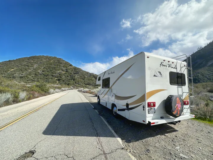 Four Winds RV rental with a Bigfoot spare tire cover, parked on a winding mountain road under a blue sky.. Thor Four Winds 2019