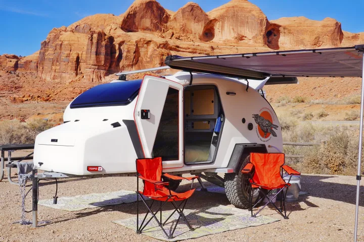 A white teardrop RV rental, perfect for outdoor adventure, is set up with an awning and chairs in front of red rock cliffs.. Escapod Escapod Trailer 2023