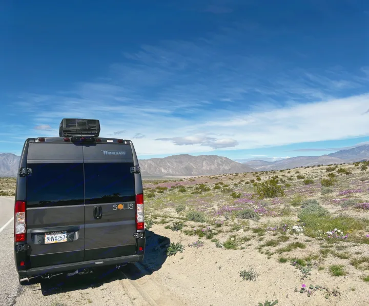 A dark grey Winnebago Solis RV rental is parked, facing a vibrant desert full of purple wildflowers and distant mountains.. Winnebago Solis 2023