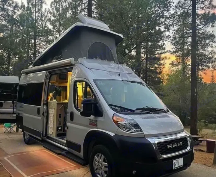 Ready for adventure: a light gray RV rental with an open pop-top & bright interior, parked in a peaceful pine forest at sunset.. Thor Sequence 2021