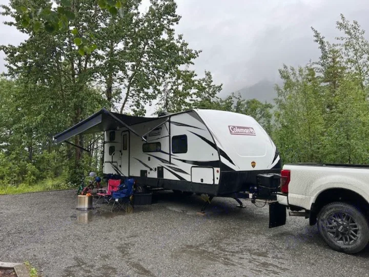 An RV rental setup at a wet campground. A white travel trailer is connected to a truck, with chairs and a fire pit under an awning.. Dutchmen Coleman Light 2021