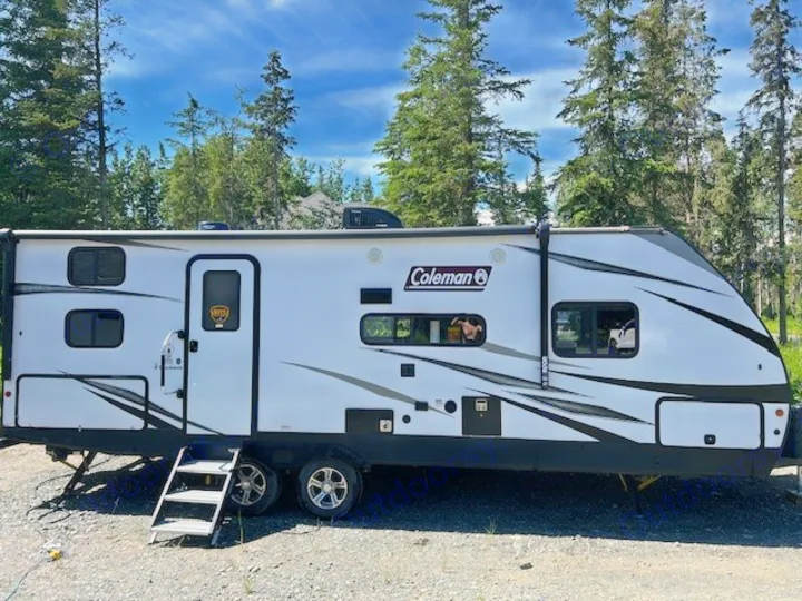 White Coleman RV rental parked on gravel with steps down, nestled among pine trees under blue skies. Perfect for a camping trip.. Dutchmen Coleman Light 2021