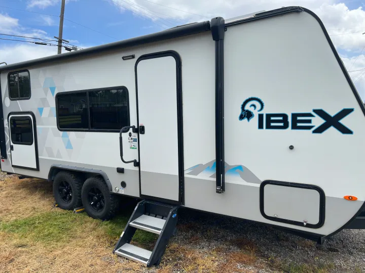 Side view of an Ibex RV rental travel trailer, white and grey with blue mountain graphics, black wheels, and steps.. Forest River Ibex 2021