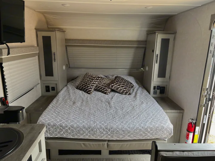RV rental interior: A cozy bedroom with patterned bed, light wood storage, TV, and window with blinds.. Forest River Ibex 2021