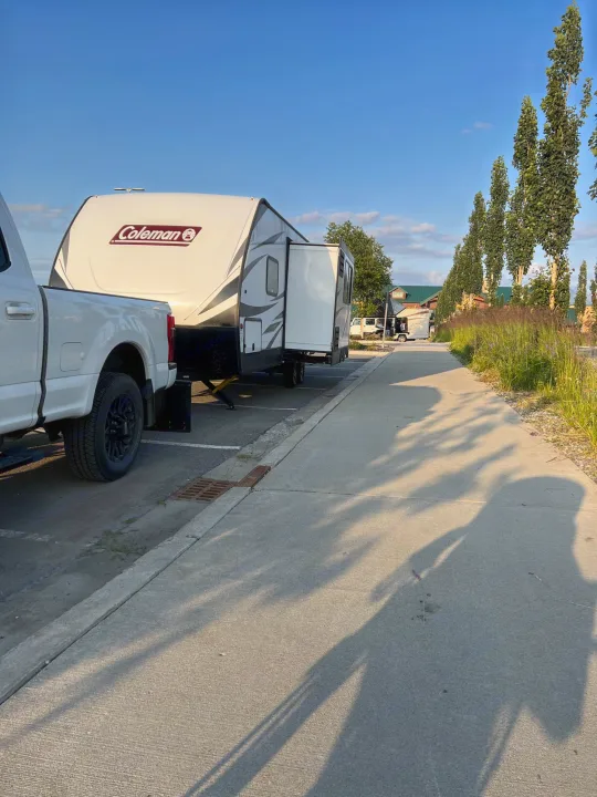 White truck hitched to a Coleman RV rental, parked in a sunny lot. Long shadows stretch across the pavement.. Dutchmen Coleman Light 2021