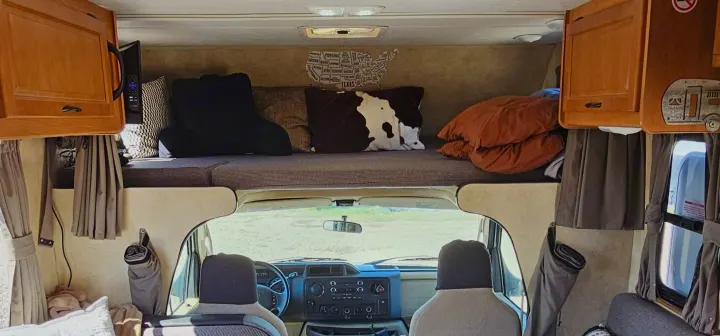 Cozy RV rental interior: overhead bunk with pillows, cabinets, and front cab view. Perfect for travel & camping.. Thor Majestic 2014
