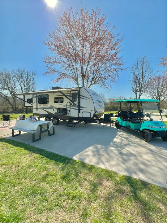 Sunny RV rental campsite featuring a travel trailer, picnic table with settings, and turquoise golf cart.. Keystone RV Outback Ultra-Lite 2018