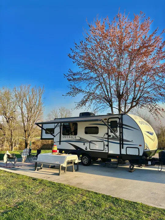 This RV rental is perfectly set up at a sunny campsite by a river, featuring a picnic area and blooming trees.. Keystone RV Outback Ultra-Lite 2018