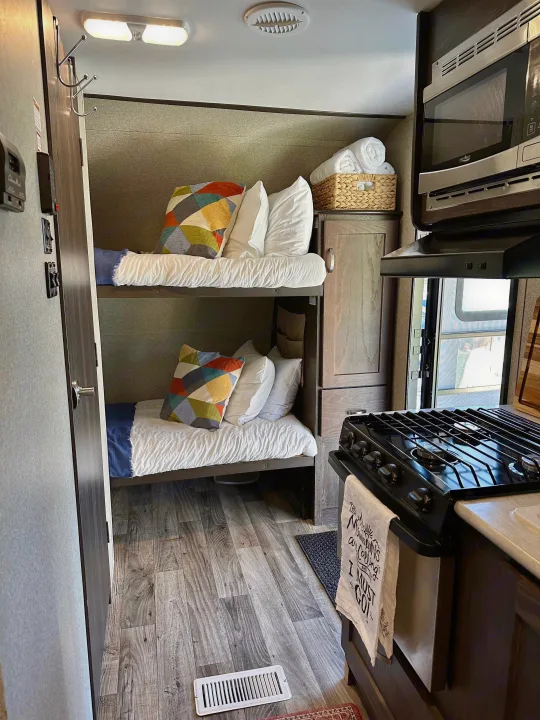 Cozy RV rental interior featuring bunk beds, a compact kitchen with stove and microwave, and wood-look flooring. Perfect for family adventures.. Keystone RV Outback Ultra-Lite 2018