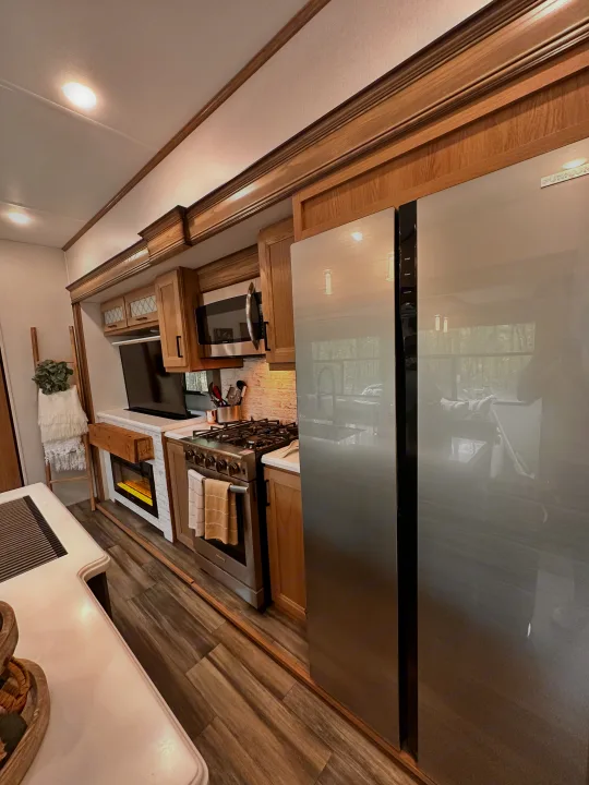 A modern RV rental's kitchen and living area, featuring a full fridge, gas range, electric fireplace, and elegant wood cabinetry.. Primetime Sanibel 3602WB 2023