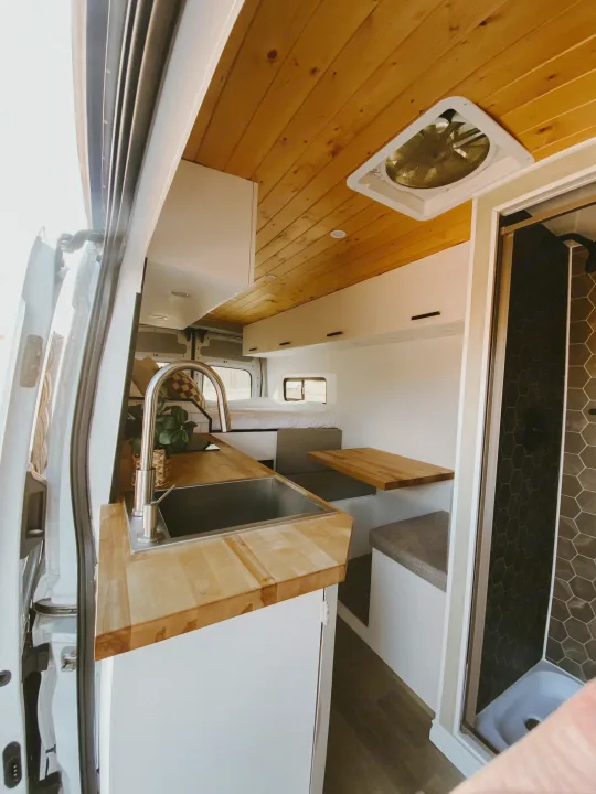 Explore this stylish RV rental interior, showcasing a modern kitchen, cozy bed, dining area, and hexagon-tiled shower.. Ford Transit 2020