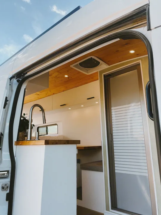 View inside a modern RV rental's bright interior: kitchen, wood ceilings, bed, and frosted sliding door.. Ford Transit 2020