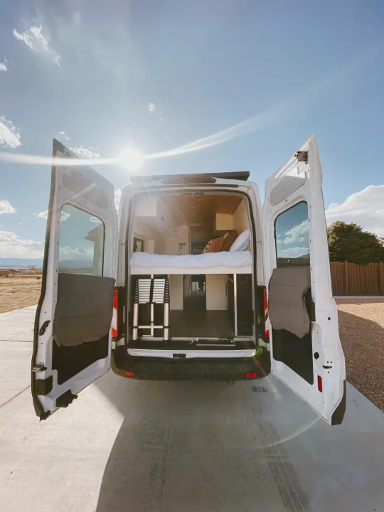 Sun-drenched white camper van, ideal RV rental. Rear doors open to reveal a cozy bed & storage, ready for van life adventures.. Ford Transit 2020