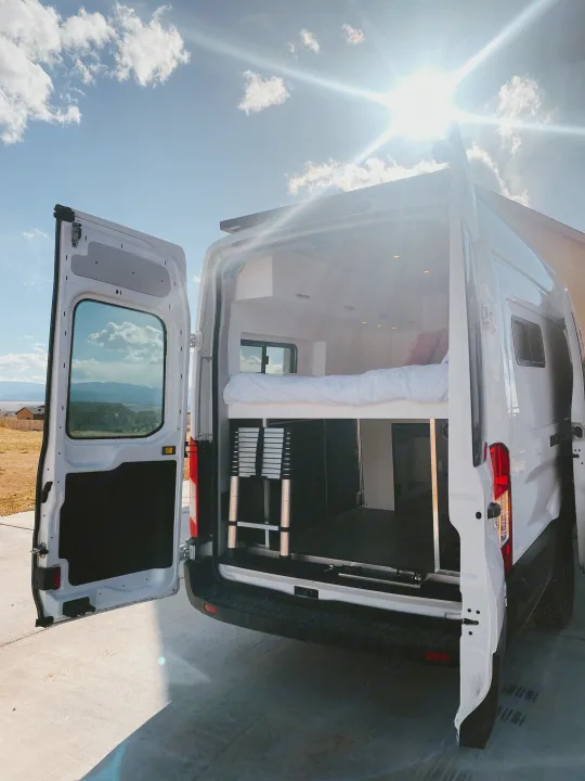 Explore our RV rental camper van's interior! Rear doors open to reveal a comfy bed, storage, and ladder, perfect for sunny day travel.. Ford Transit 2020