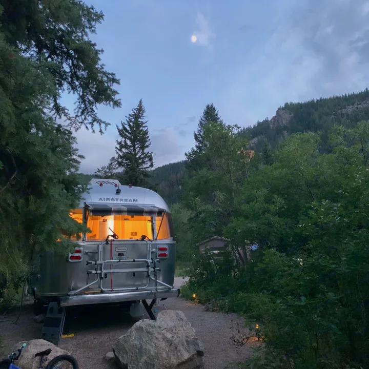 A cozy Airstream RV rental glowing at dusk, nestled in mountains under a bright moon.. Airstream Globetrotter 2020