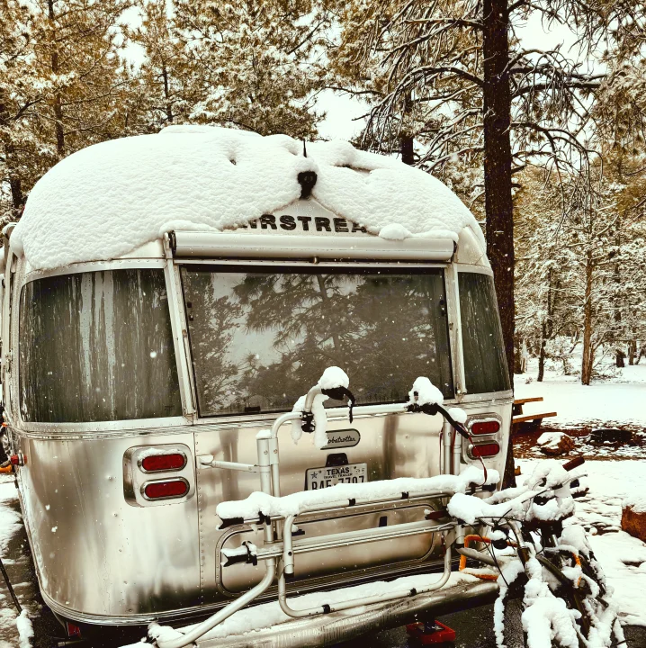 Snow-covered Airstream RV with bikes on rack, parked among snowy pines. Discover winter camping with this amazing rv rental.. Airstream Globetrotter 2020