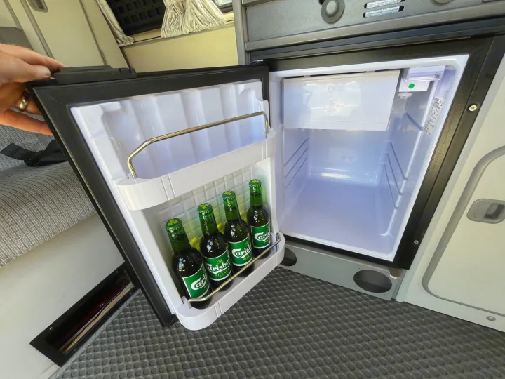 A person's hand holds open a compact fridge in an RV rental, revealing four green Carlsberg beer bottles inside. Great camper amenity.. Volkswagen Westfalia 1987