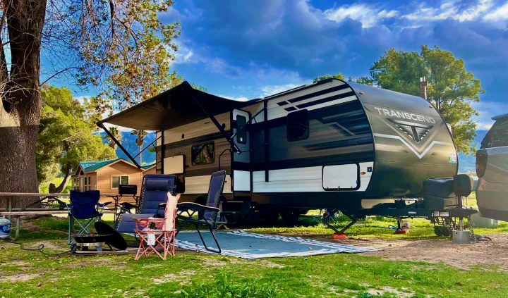 Beautiful campsite with an RV rental travel trailer set up. Awning out, chairs, and fire pit on green grass under blue skies.. Grand Design Transcend Xplor 2022