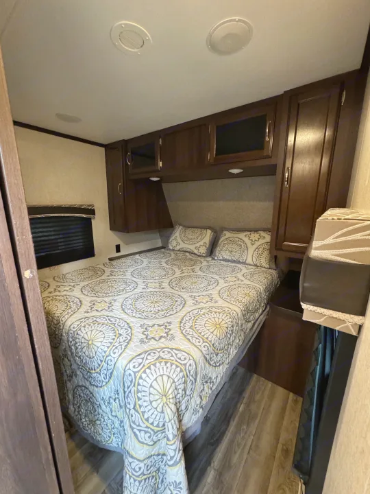Cozy RV rental bedroom featuring a queen bed with a patterned comforter, dark wood overhead storage, and a window.. Jayco Jay Flight 2018