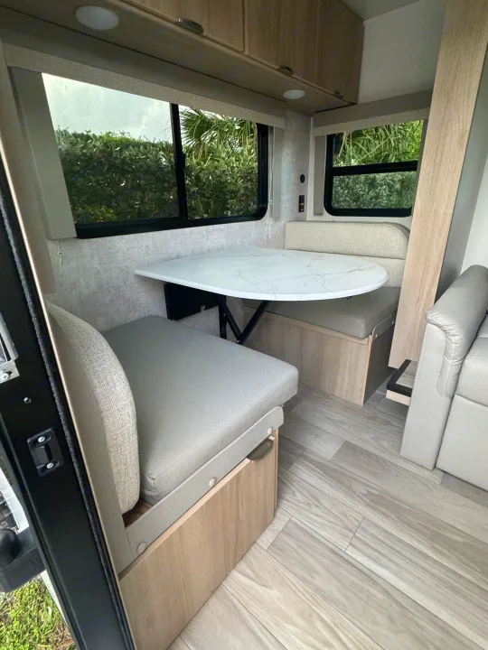 Modern RV rental interior: Dinette area with a white marble-look table, light bench seating, and large windows overlooking greenery.. Minnie Micro Minnie 2024