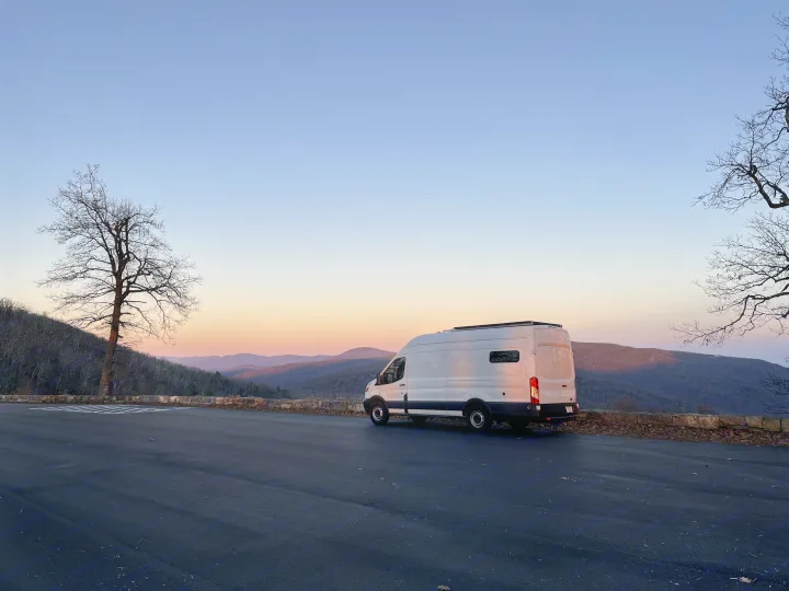 A white rv rental camper van overlooks a tranquil mountain range during a soft sunset, framed by bare trees on a paved lot.. Ford Transit 2015