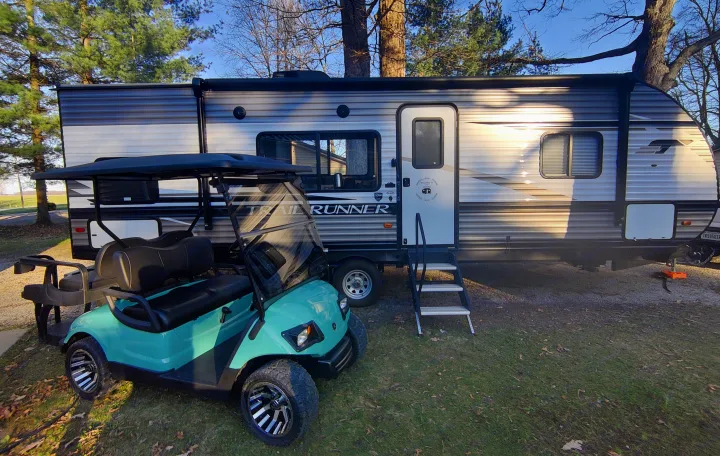 A modern RV rental, a "Trail Runner," is parked alongside a teal golf cart, suggesting a relaxed camping experience.. Heartland RVs Trailrunner 2021