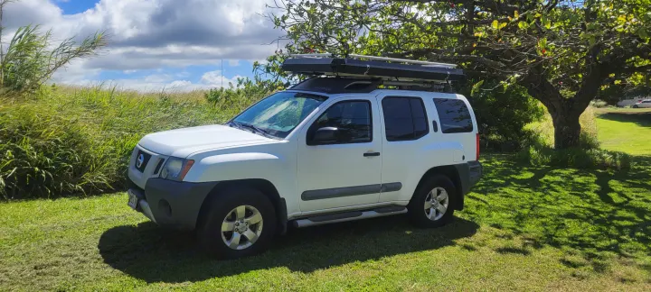 White Nissan Xterra SUV with a rooftop tent parked on grass, offering a compact outdoor vehicle for RV rental adventures.. Nissan Xterra 2012