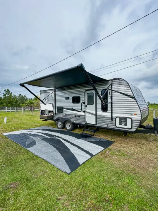 A light gray travel trailer RV rental, with an extended black awning and patterned rug on green grass, set up for camping.. Jayco Jay Flight SLX 2021