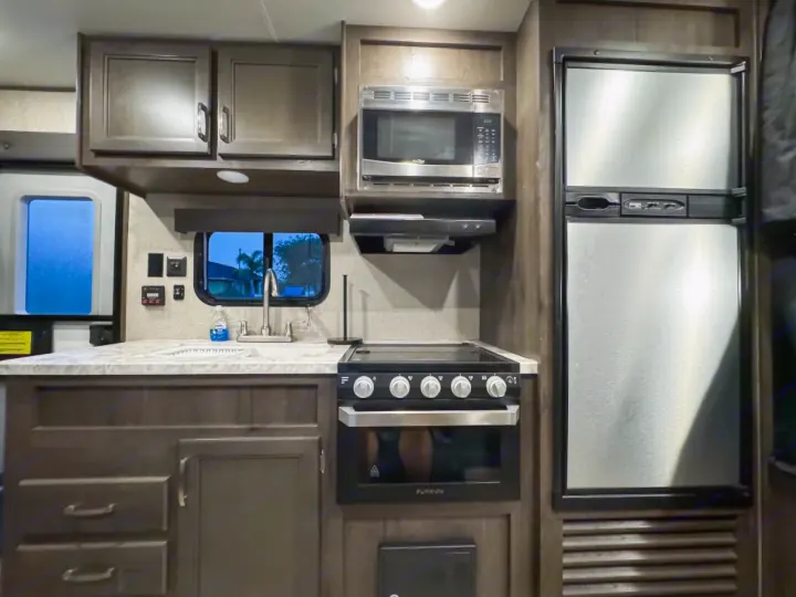 Modern RV rental kitchen with dark cabinets, stainless appliances: microwave, oven, stove, fridge, and sink. Ready for travel!. Jayco Jay Flight SLX 2021