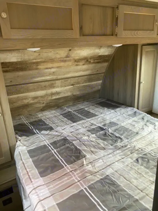 RV rental interior: A compact bedroom with a plaid bed, rustic wood headboard, and overhead storage cabinets.. Gulf Stream Gulf Breeze 2023