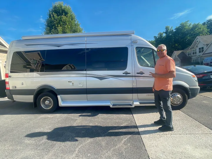 Smiling man gives peace sign beside a sleek, silver Pleasure-Way Plateau Sprinter RV, ready for an RV rental journey.. Pleasure-Way Pleasure-Way Class B 2013