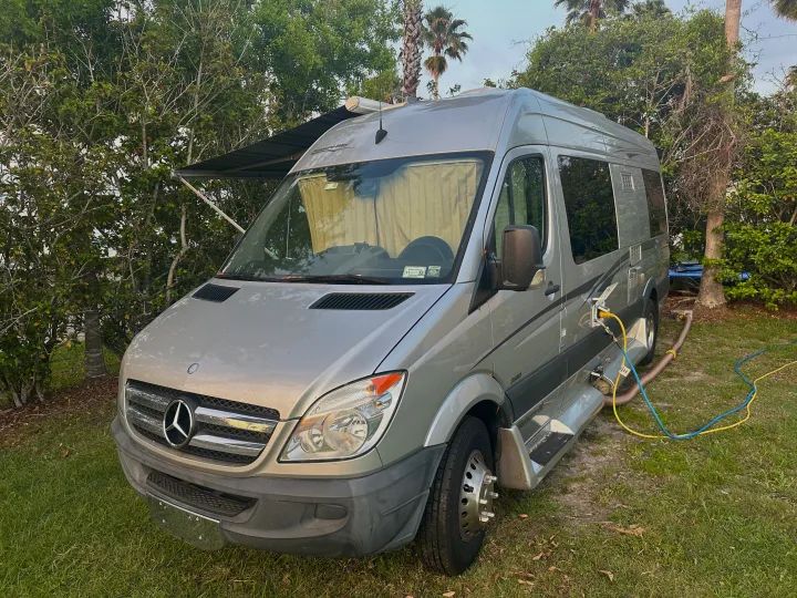 Silver Mercedes Sprinter RV rental parked on grass with hookups connected, awning extended.. Pleasure-Way Pleasure-Way Class B 2013