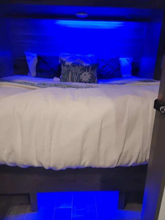 Cozy RV rental bedroom features a bed with blue LED lighting illuminating the headboard and floor. White bedding with decorative pillows creates a relaxing ambiance.. Crossroads RV Sunset Trail Super Lite 2021