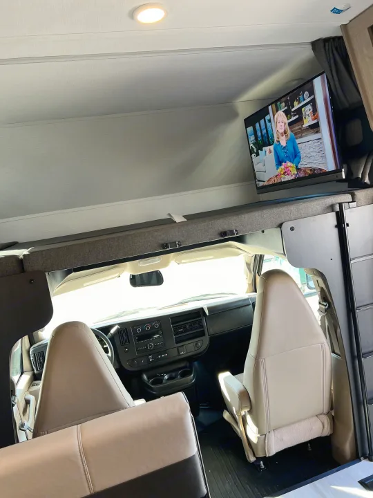 Interior of an RV rental, showing the front cabin area with driver and passenger seats, dashboard, and an overhead TV.. Forest River Sunseeker 2025