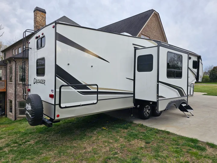 Large white RV rental, a travel trailer, with a rear ladder and extended slide-out, parked by a brick house.. Crusader Lite 2022