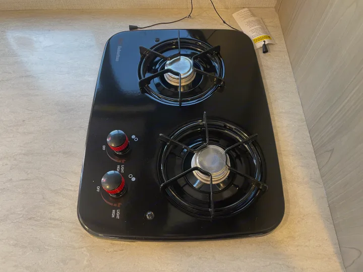 Black Suburban 2-burner gas stove inside an RV rental. Visible are burner grates, control knobs for 'LIGHT' and 'HIGH', and a warning tag.. Keystone Springdale Classic Mini 2024