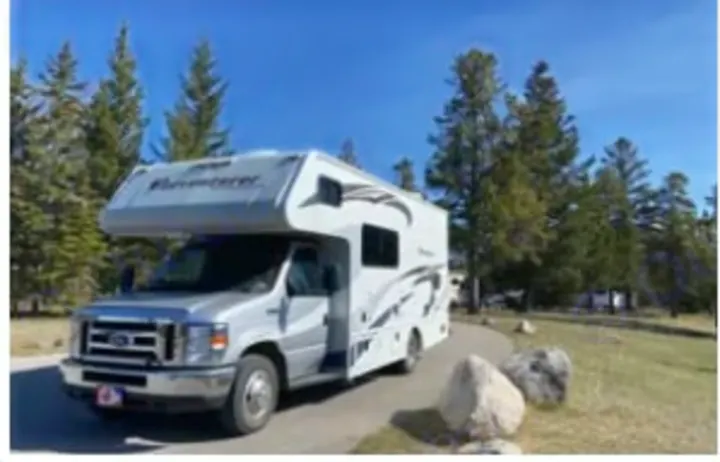 A white & silver rv rental parked on a sunny paved spot, surrounded by tall pine trees. Perfect for your outdoor travel & camping.. Adventurer Adventurer Motorhome 2019