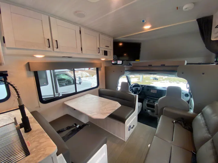 Bright RV rental interior featuring a functional kitchen, dinette seating with seatbelts, and a view into the cab.. East to West Entrada 2025