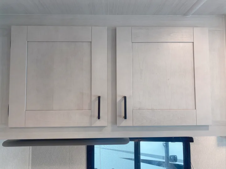 Light wood overhead cabinets with black handles in an RV rental interior, above an overhead fixture and window.. East to West Entrada 2025