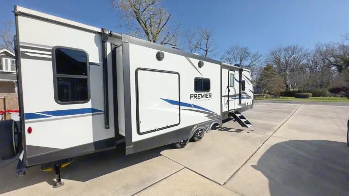 This sleek white travel trailer RV rental, featuring blue accents and an extended slide-out, is parked on a driveway on a clear day.. Keystone RV Bullet Premier Ultra Lite 2022