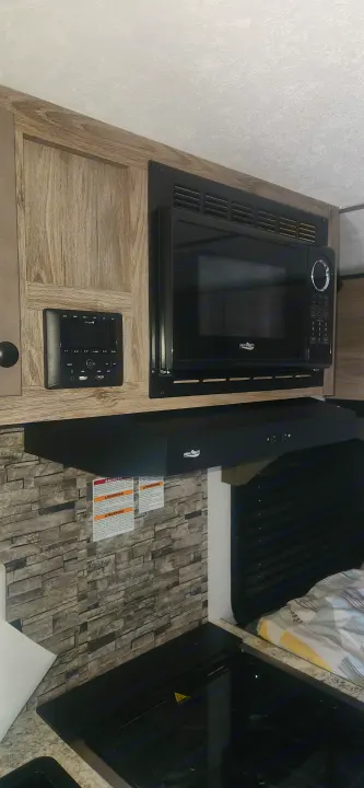 RV rental kitchen: High Pointe microwave, range hood, and electric cooktop above stone backsplash & wood cabinets.. Sunset Park RV Sun Lite 2024