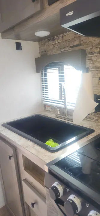 This RV rental kitchen features a black sink, faucet, cooktop, stone backsplash, and cabinets, offering functional living space.. Sunset Park RV Sun Lite 2024
