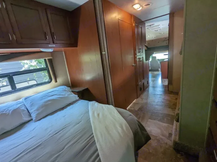 RV rental interior: a comfy bedroom with a window, dark wood cabinets, and a tiled hallway leading to the front cab.. Jayco Redhawk 2015