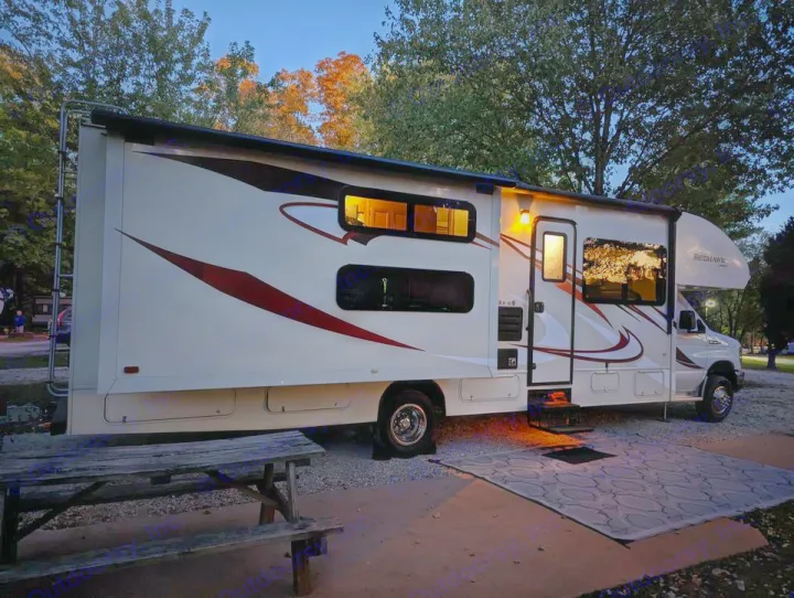 This inviting white RV rental is set up at a dusk campsite. Interior lights glow, illuminating the porch area and outdoor mat.. Jayco Redhawk 2015