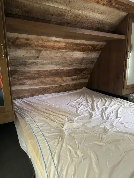 RV rental bedroom interior, featuring wood-paneled walls, an overhead shelf, and a bed with a white sheet.. Keystone RV Passport SL 2021
