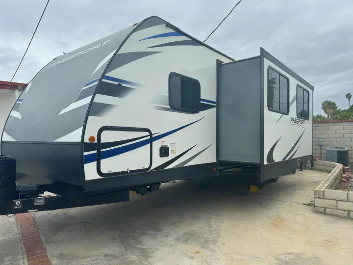 A white, gray, and blue Passport travel trailer **rv rental** with its side slide-out extended, parked on a concrete driveway.. Keystone RV Passport SL 2021